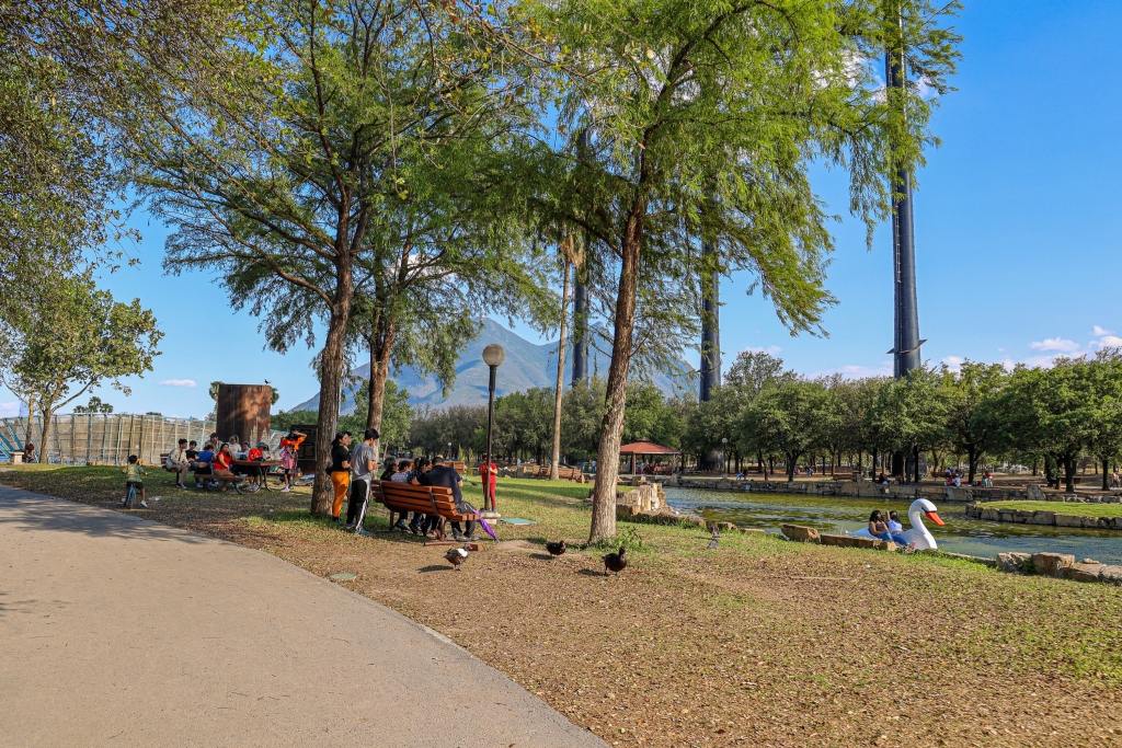 Familias descansan en el Ã¡rea verde junto a la laguna, con patos, lancha de cisne y chimeneas industriales.
